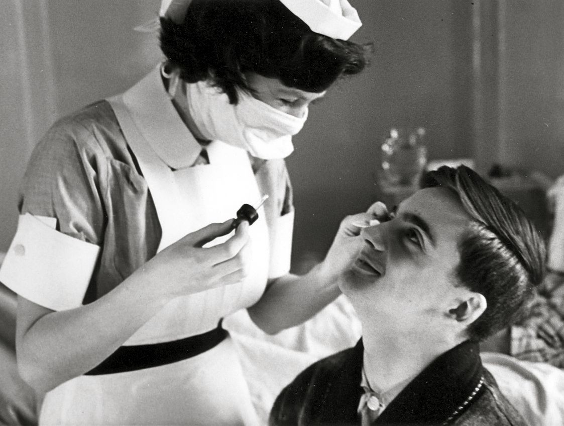 1950s image of a masked nurse placing eye drops into a patient's eyes