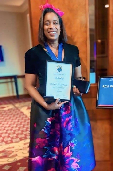 Roxanne Crosby Nwaobi, wearing a beautiful and colourful dress, holding her award and smiling
