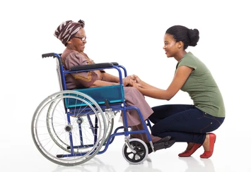 Two people facing each other holding hands, one sitting in a wheelchair and the other crouched holding their hands