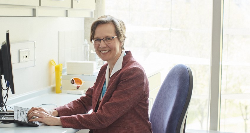 Photo of smiling consultant looking to camera, sitting at a desk in a consultation room